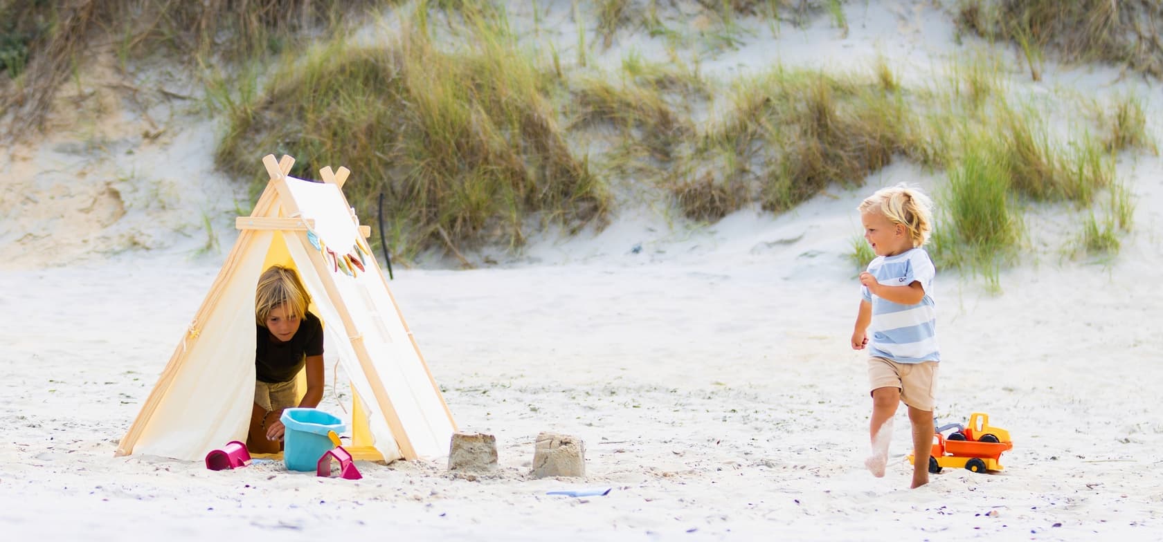 Twee kinderen spelen op het zandstrand van Groede – één zit in een klein tentje met kleurrijke vlaggetjes, de ander loopt met een speelgoedtruck door het zand.