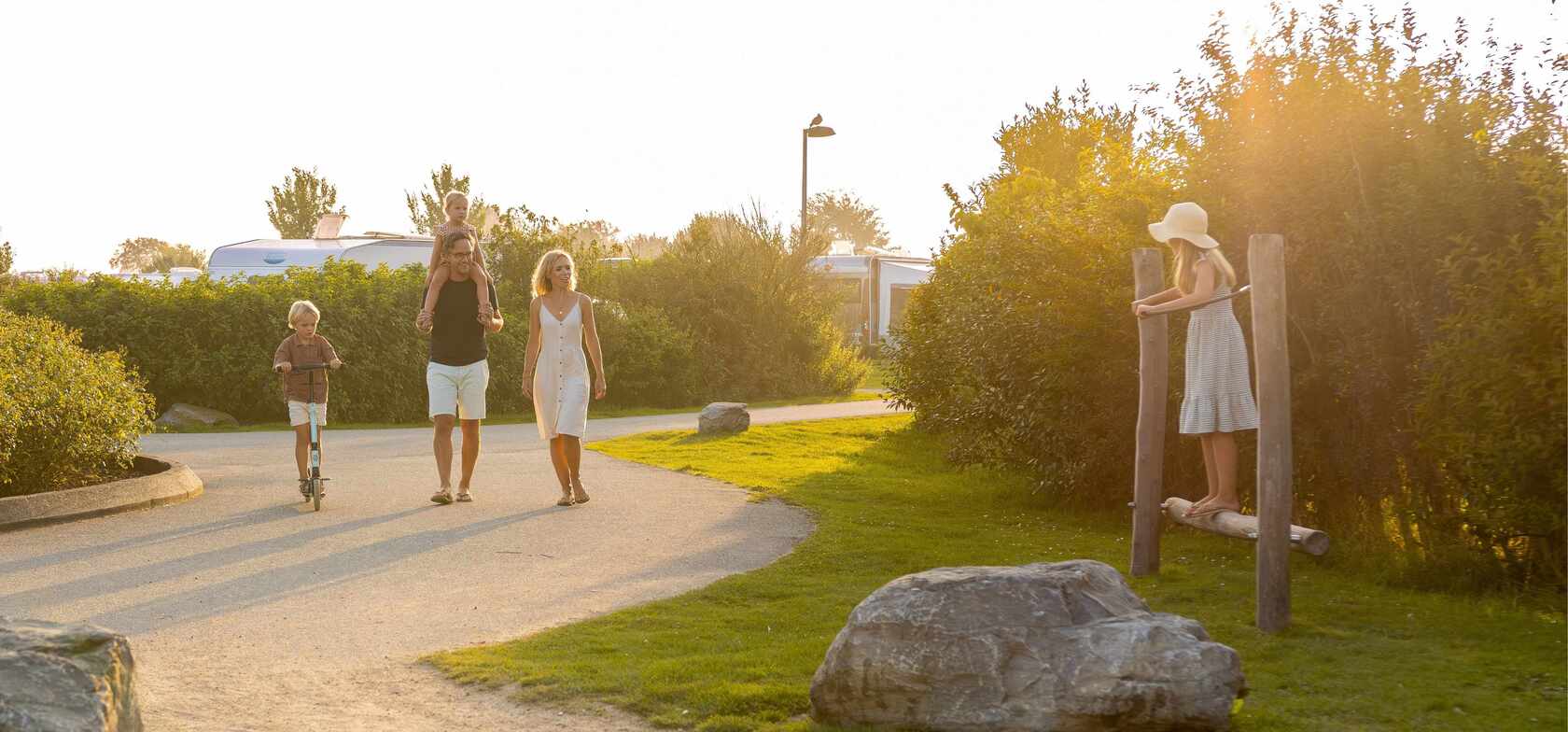 Eine Familie erkundet gemeinsam die Umgebung des Strandcamping Groede – mit einem Kind auf einem Roller, einem Erwachsenen mit einem Kind auf den Schultern und einem weiteren Kind, das auf Holzpfählen spielt, umgeben von Natur und Campingfahrzeugen.