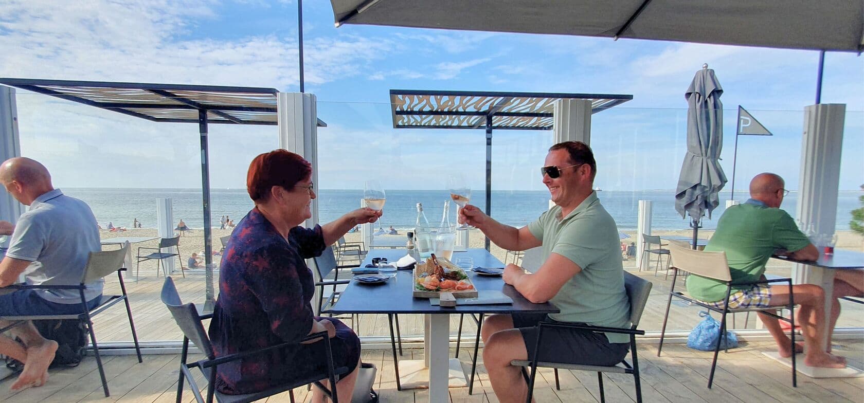 Zwei Personen stoßen mit Gläsern Weißwein auf einer Strandterrasse des Strandcamping Groede an, mit Blick auf das Meer und andere Gäste, die ihr Essen genießen
