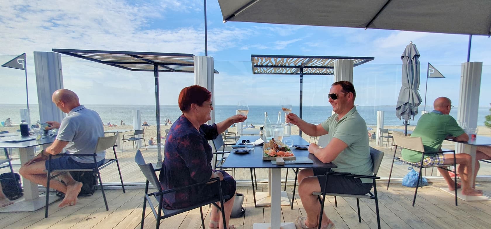 Twee mensen proosten met witte wijn op een strandterras van Strandcamping Groede, met uitzicht op zee en andere gasten die van hun maaltijd genieten.