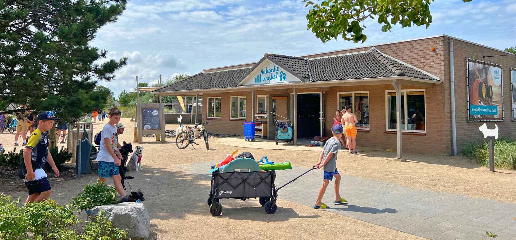 Der Eingang zum Ferienladen auf dem Strandcamping Groede mit dem Schild ‚Vakantiewinkel‘, umgeben von Fahrrädern, Bäumen und Sommergästen – darunter ein Kind mit einem Bollerwagen voller Strandspielzeug.