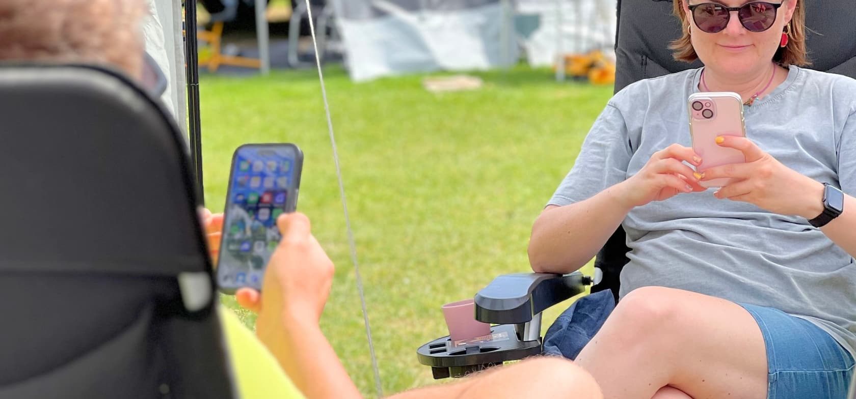 Twee mensen zitten buiten op stoelen en gebruiken hun smartphones – met zichtbare app-iconen en een roze hoesje – omgeven door gras en zomerse sfeer.