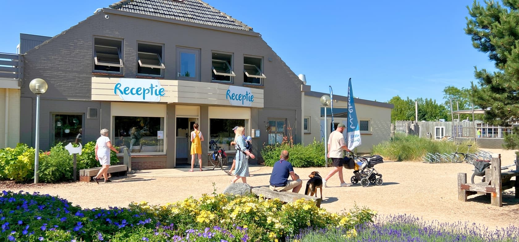 Das Rezeptionsgebäude des Strandcamping Groede mit dem Schild „Rezeption“, davor eine Familie mit Kinderwagen und Hund – umgeben von Blumen und blauem Himmel.
