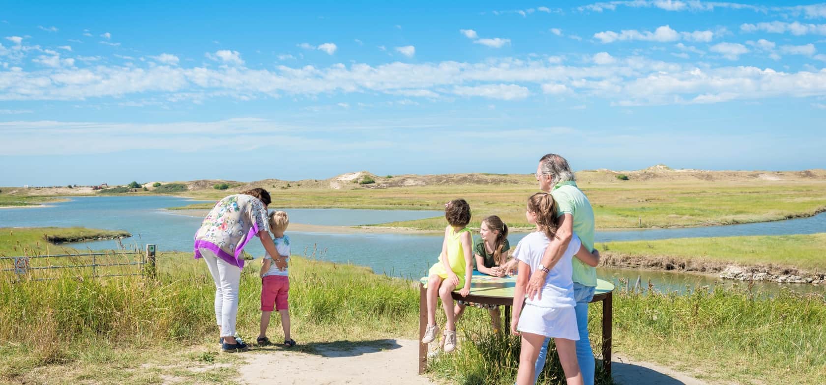 Een familie geniet van de natuur bij het water – omgeven door groene weiden, een rivier en een blauwe lucht met wolken. Samen ontdekken ze de omgeving.