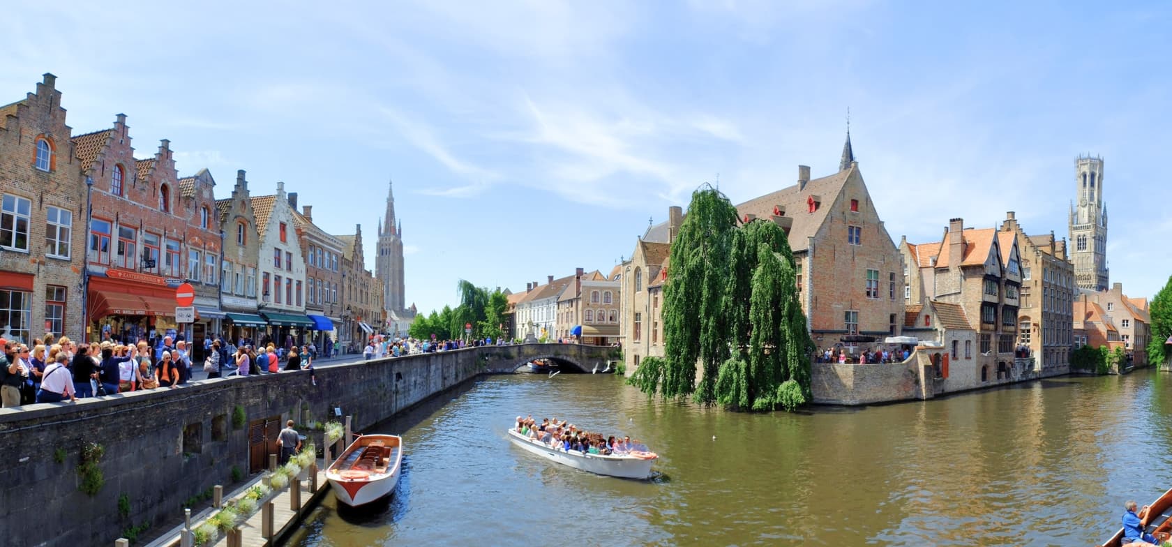 Een schilderachtig uitzicht op de grachten van Brugge in België – met historische gebouwen, wandelaars langs de oever, rondvaartboten en een kerktoren op de achtergrond.