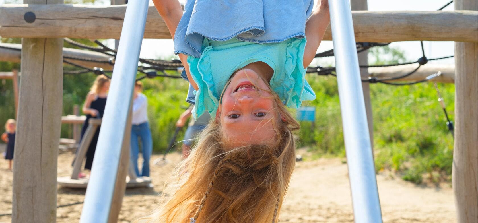 Ein Kind hängt kopfüber an einem Spielgerät mit den Beinen über einer Stange, umgeben von anderen Kindern, Erwachsenen, Sand und viel Grün – ein lebendiger Ort für Spiel und Bewegung.