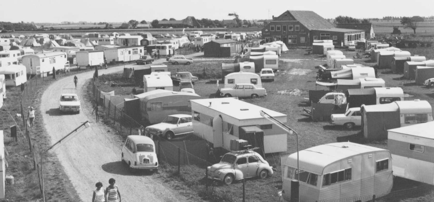 Zwart-witfoto van de vroegere camping met caravans, auto's en een gebouw op de achtergrond – een blik op de geschiedenis van Strandcamping Groede.