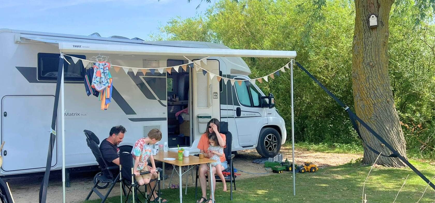 Eine Familie sitzt unter der Markise ihres Wohnmobils auf dem Strandcamping Groede, umgeben von grüner Natur – ideal für einen entspannten Familienurlaub.
