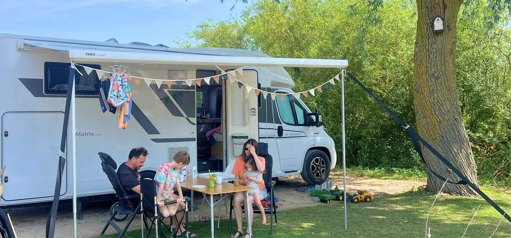 Eine Familie sitzt unter der Markise ihres Wohnmobils auf dem Strandcamping Groede, umgeben von grüner Natur – ideal für einen entspannten Familienurlaub.